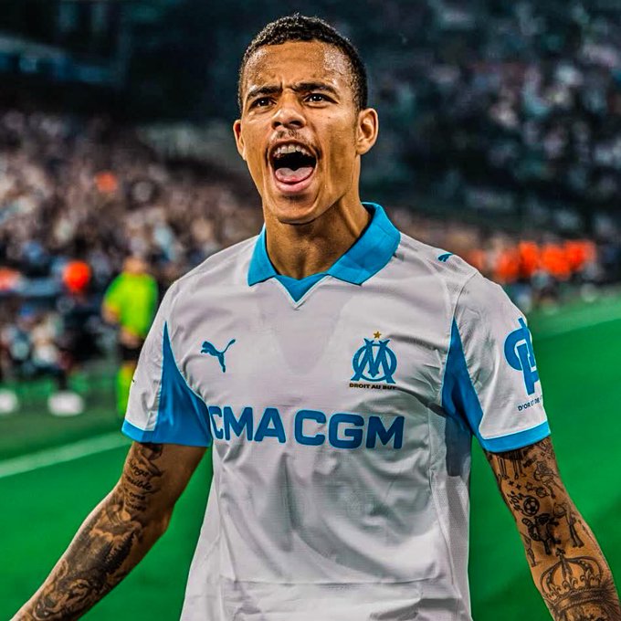 A young man with short dark hair and a focused expression stands on a green soccer field during a match, wearing a white and blue Olympique de Marseille jersey with Puma logo and sponsor patches, arms outstretched in celebration, tattoos visible on his arms, stadium crowd and orange cones in the blurred background.